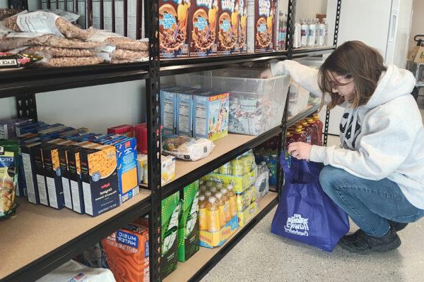 A woman wearing a gray hoodie and blue jeans is crouching down while selecting items from a shelf stocked with food and drinks. They are placing the items into a blue reusable bag with the logo